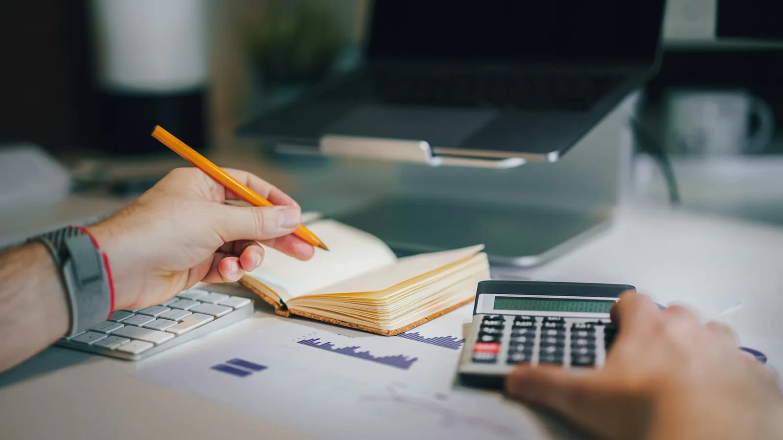 Deux mains au travail sur un bureau : un crayon au-dessus d’un carnet ouvert, une calculatrice, un graphique imprimé et un ordinateur portable.