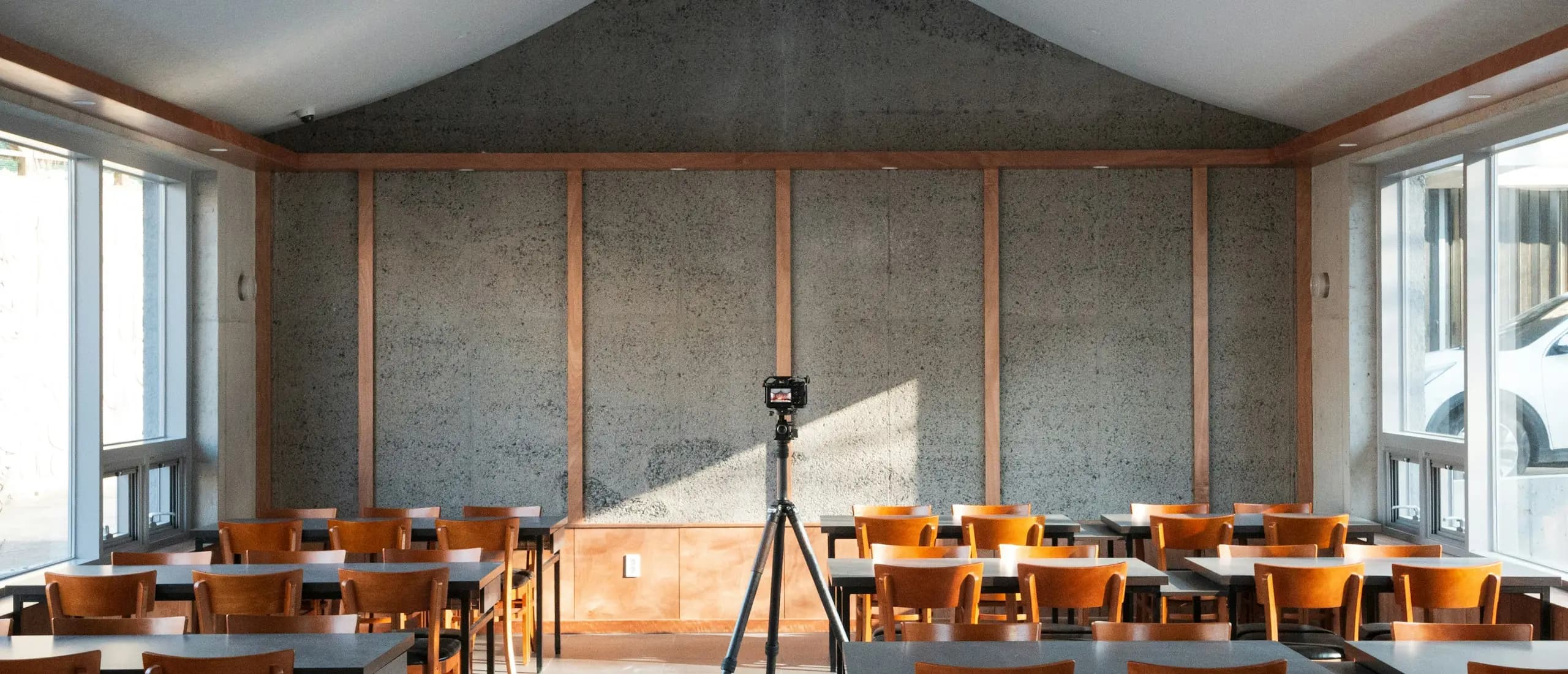 Une salle de formation vide, tables et chaises en bois face à un mur de béton, un appareil photo sur trépied au centre.