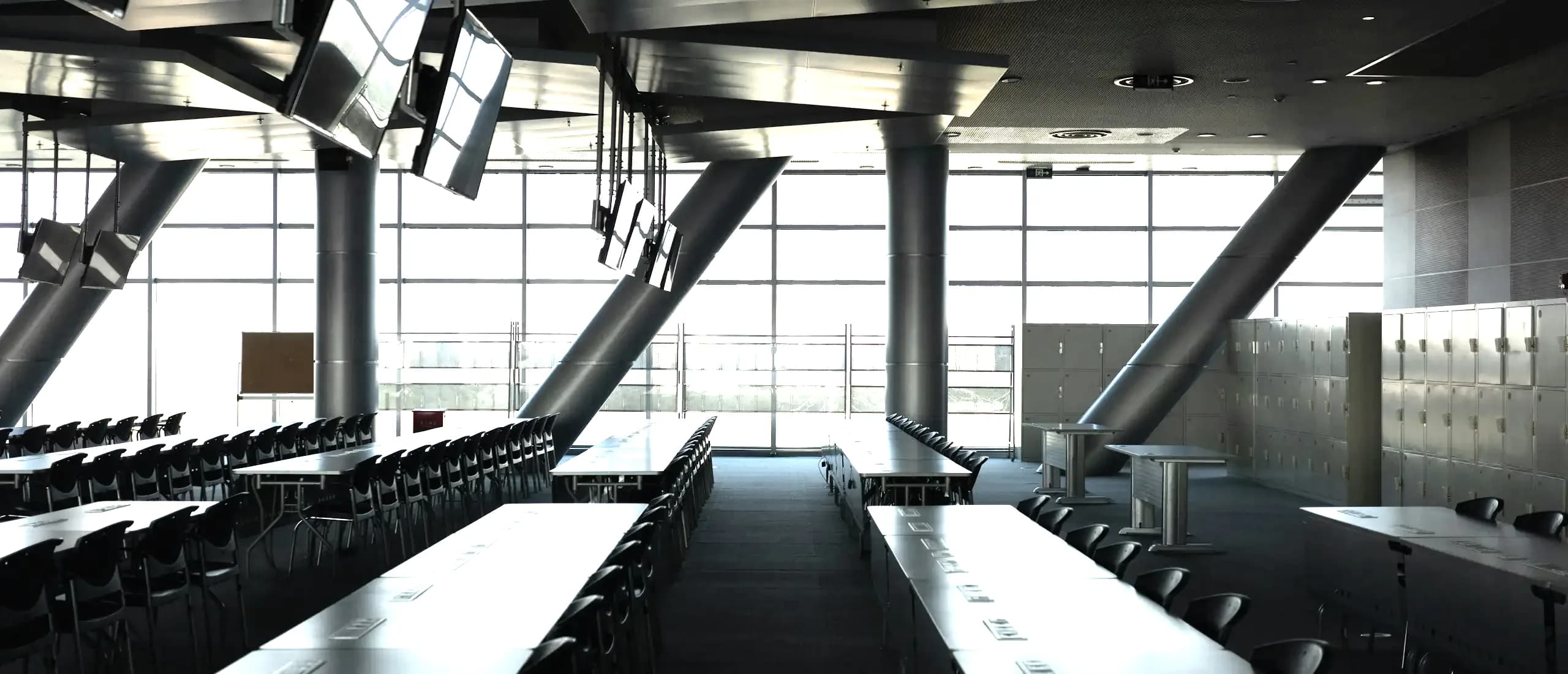 Une grande salle de travail vide : longues tables alignées, écrans suspendus et baies vitrées donnant sur la ville.