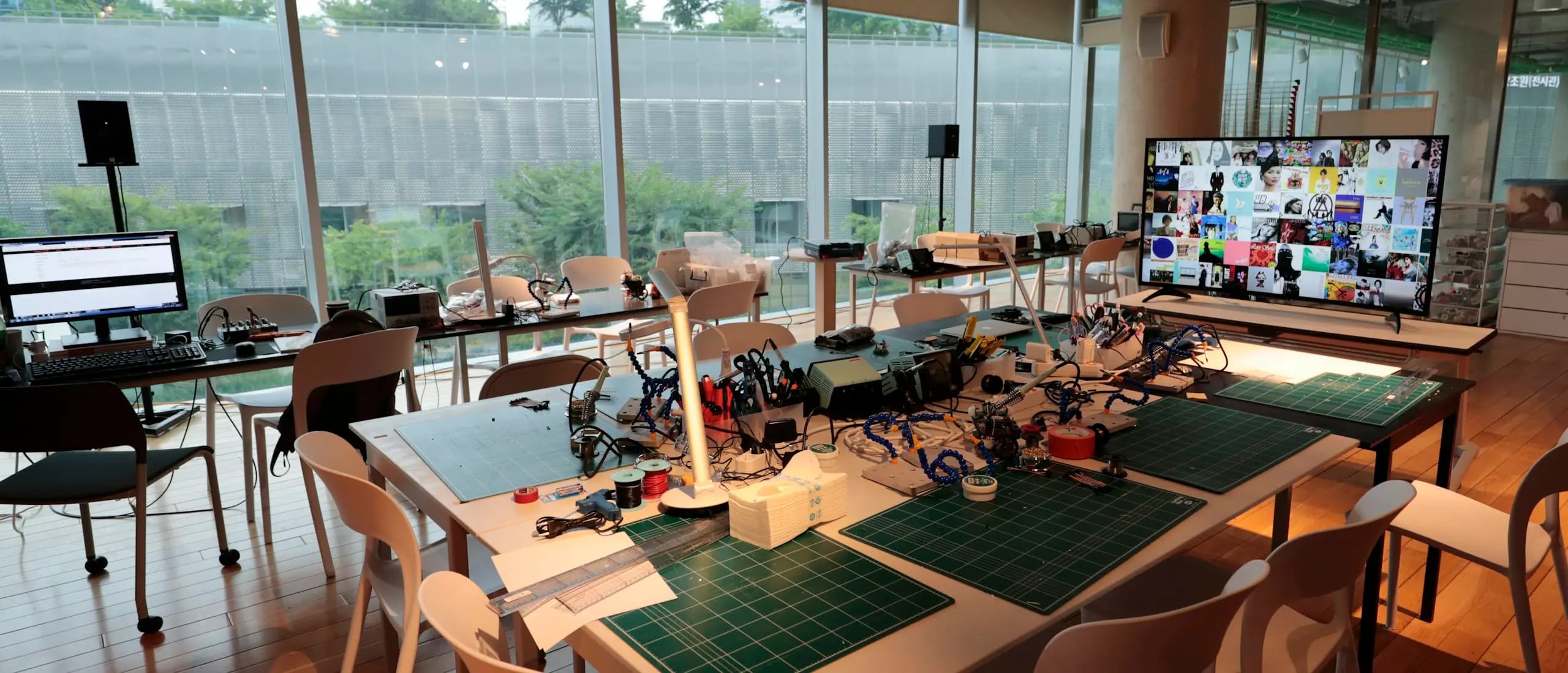 Un atelier de formation aux grandes tables claires, couvertes de tapis de découpe, d’outils et de matériel électronique, devant une baie vitrée.